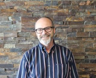 a person in a striped shirt and glasses stands in front of a stone wall